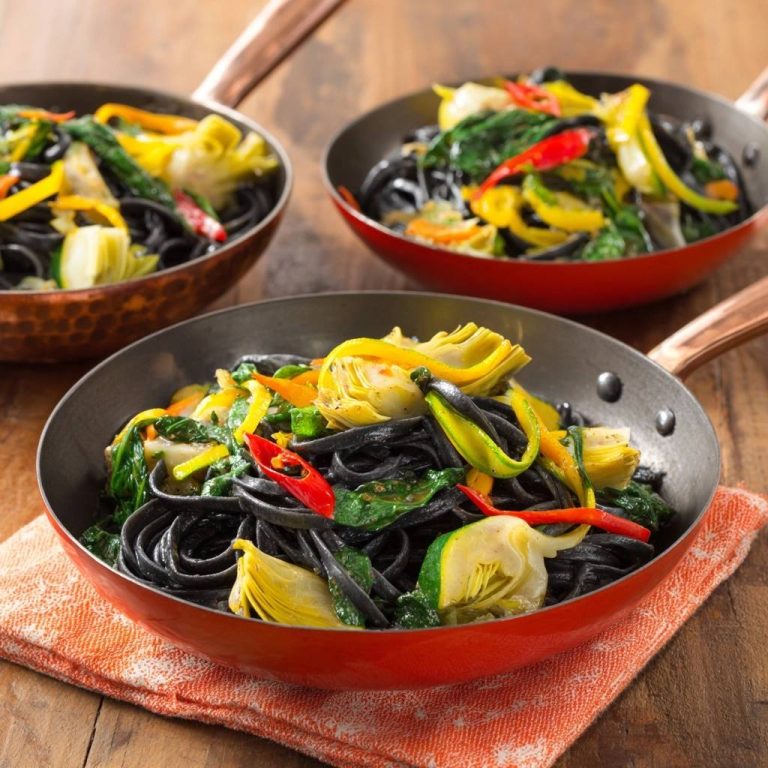 Schwarze Linguine mit Artischocken und buntem Gemüse, serviert in drei kleinen Pfannen auf einem Holztisch.